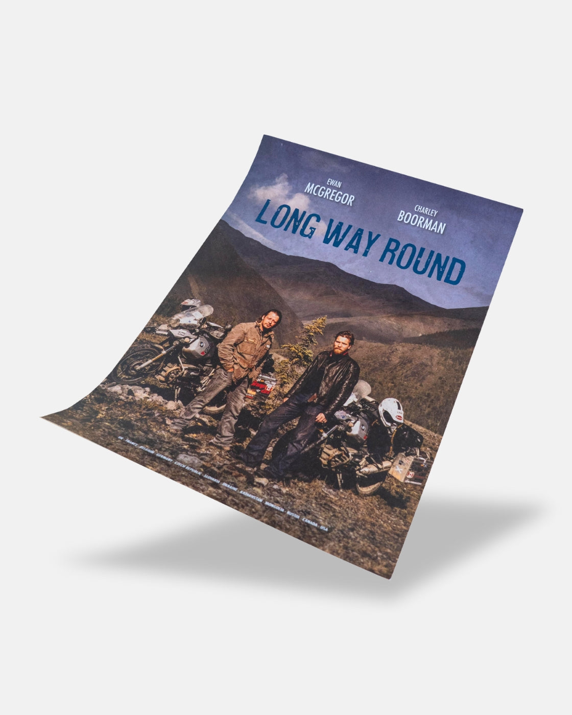 The Long Way Shop’s Long Way Round Poster features two men before motorcycles and mountain scenery under a cloudy sky, with the names Ewan McGregor and Charley Boorman at the top.