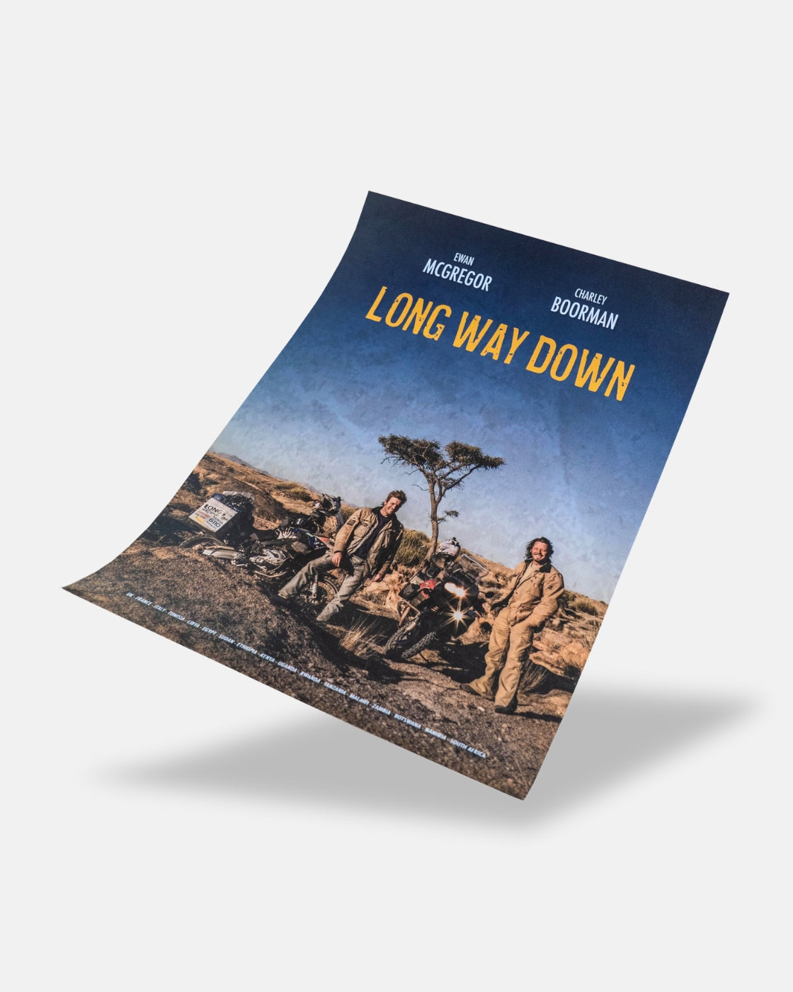 A titled Long Way Down Poster from Long Way Shop features Ewan McGregor and Charley Boorman by motorcycles in a rugged landscape with a lone tree beneath a blue sky.