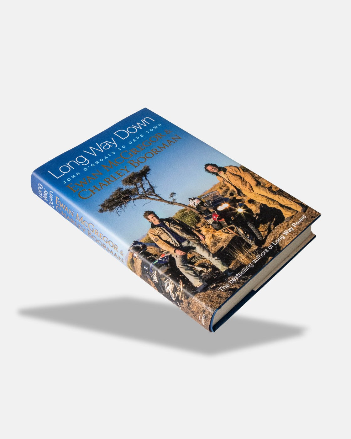 The Long Way Shop’s Long Way Down Book (Hardback) features a cover photo of Ewan McGregor and Charley Boorman in motorcycle gear beside their bikes in a rugged outdoor setting with a tree in the background.