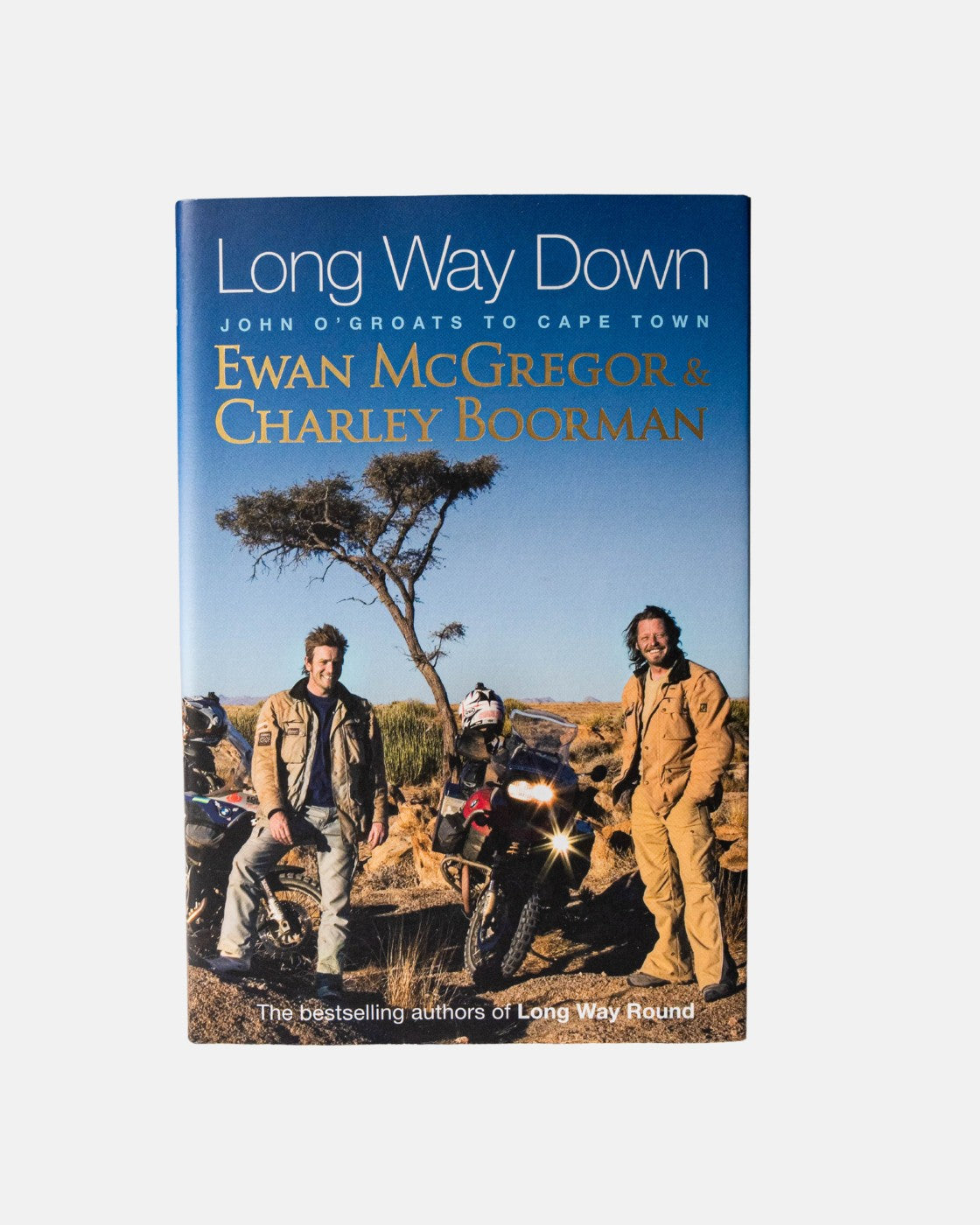 Hardback cover of Long Way Down Book from Long Way Shop, featuring Ewan McGregor and Charley Boorman with motorcycles under a tree on an African landscape beneath a blue sky.
