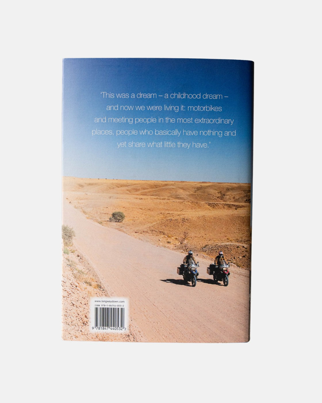 Two people ride motorcycles along a dusty road in a desert beneath a clear sky, where a quote about dreams and generosity appears. The scene is featured on the cover of the Long Way Down Book (Hardback) from Long Way Shop.