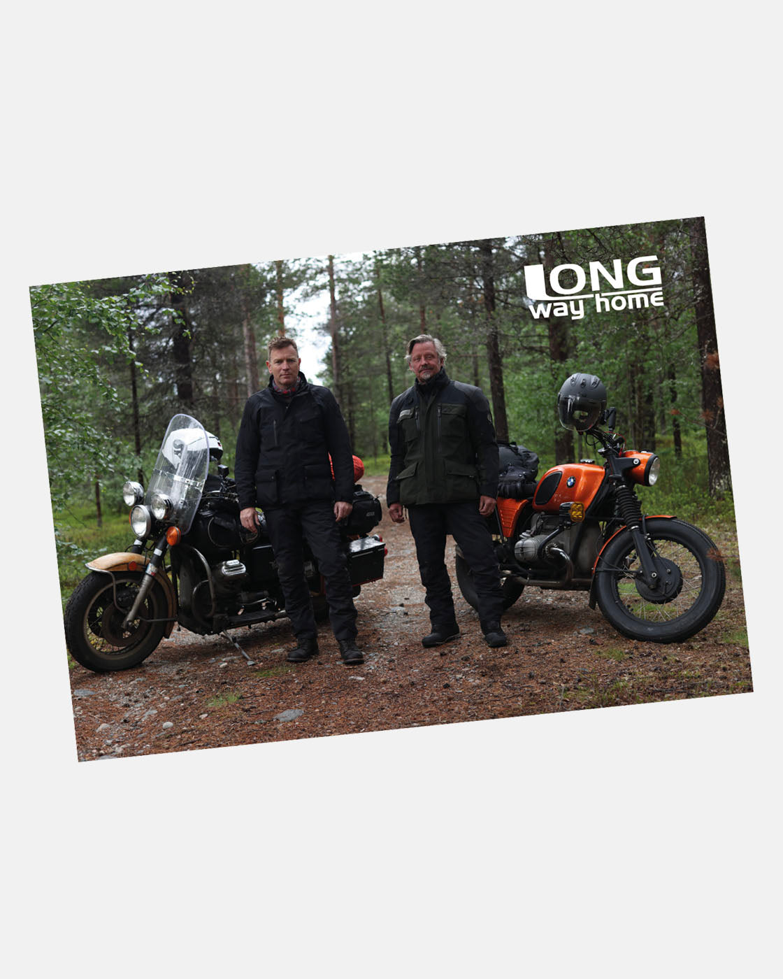 Two men in dark jackets stand by motorcycles on a forest path with trees behind them. LONG way home appears in the upper right. Available as an A2 Poster from Long Way Shop.